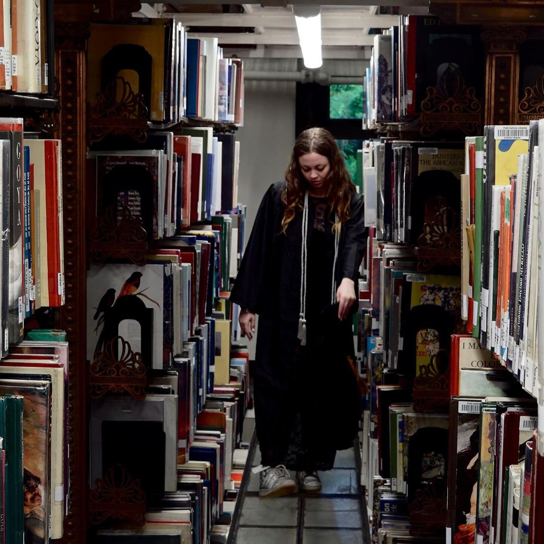 Person in a commencement gown walking down the aisle of the Pratt Institute Library.