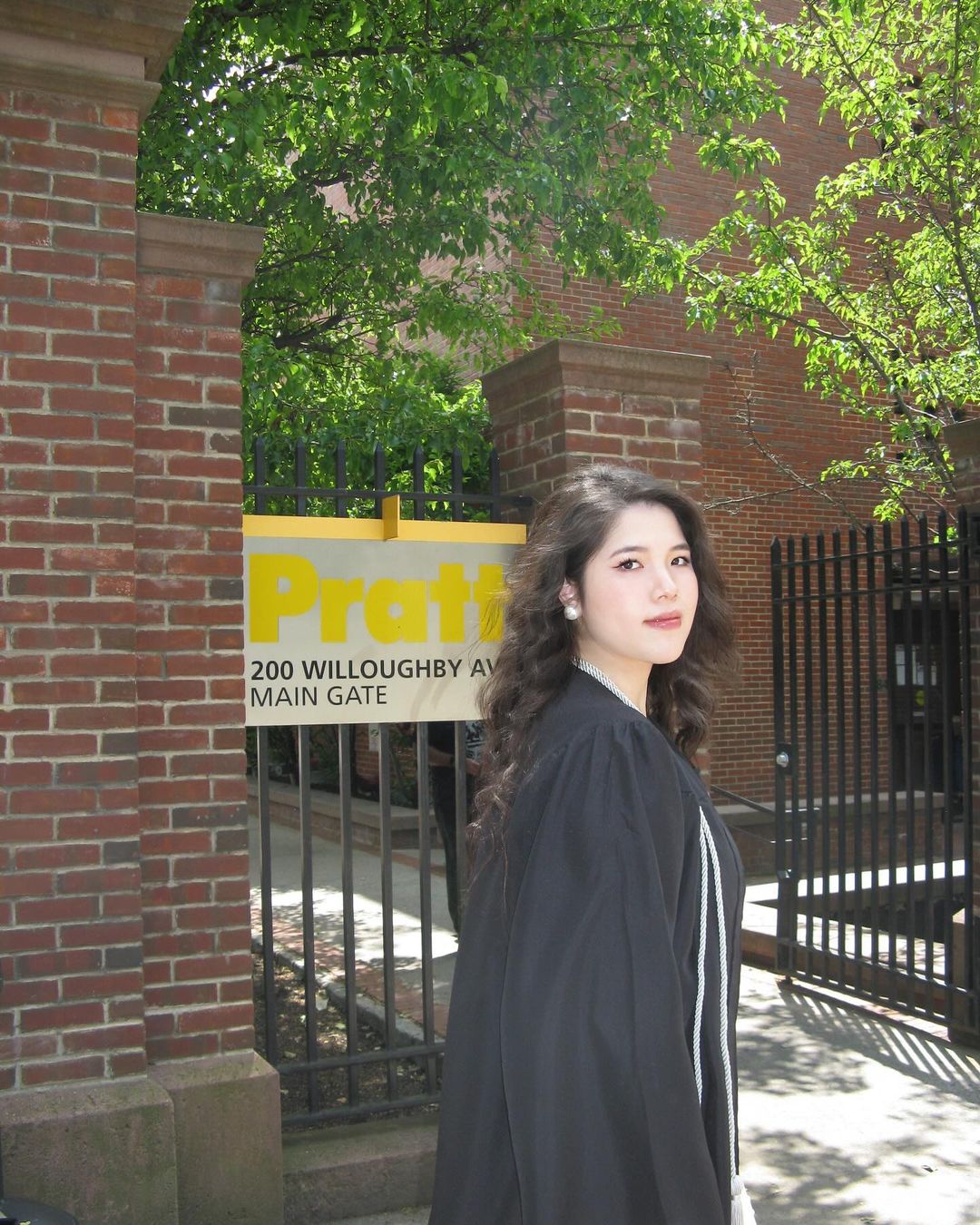 Person in their commencement gown standing in front of a Pratt sign on campus and smiling.