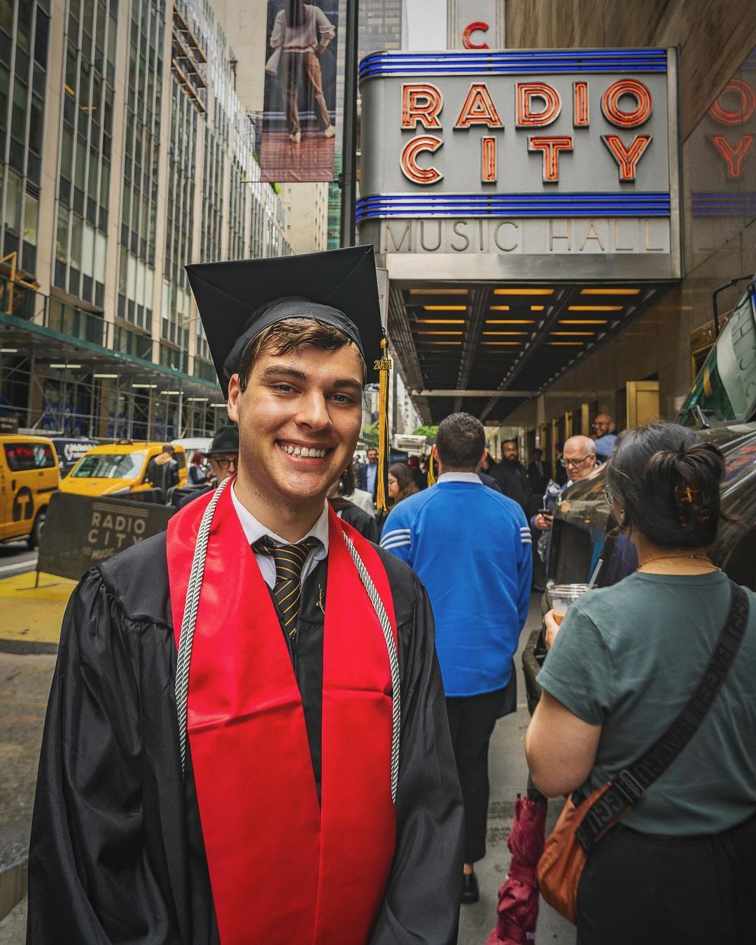 Person in a tie and commencement cap and gown standing and smiling in front of the exterior of Radio City Music Hall.