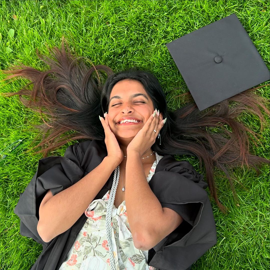 Person in a floral dress and their commencement gown lying in the grass and smiling with both hands on their cheeks.