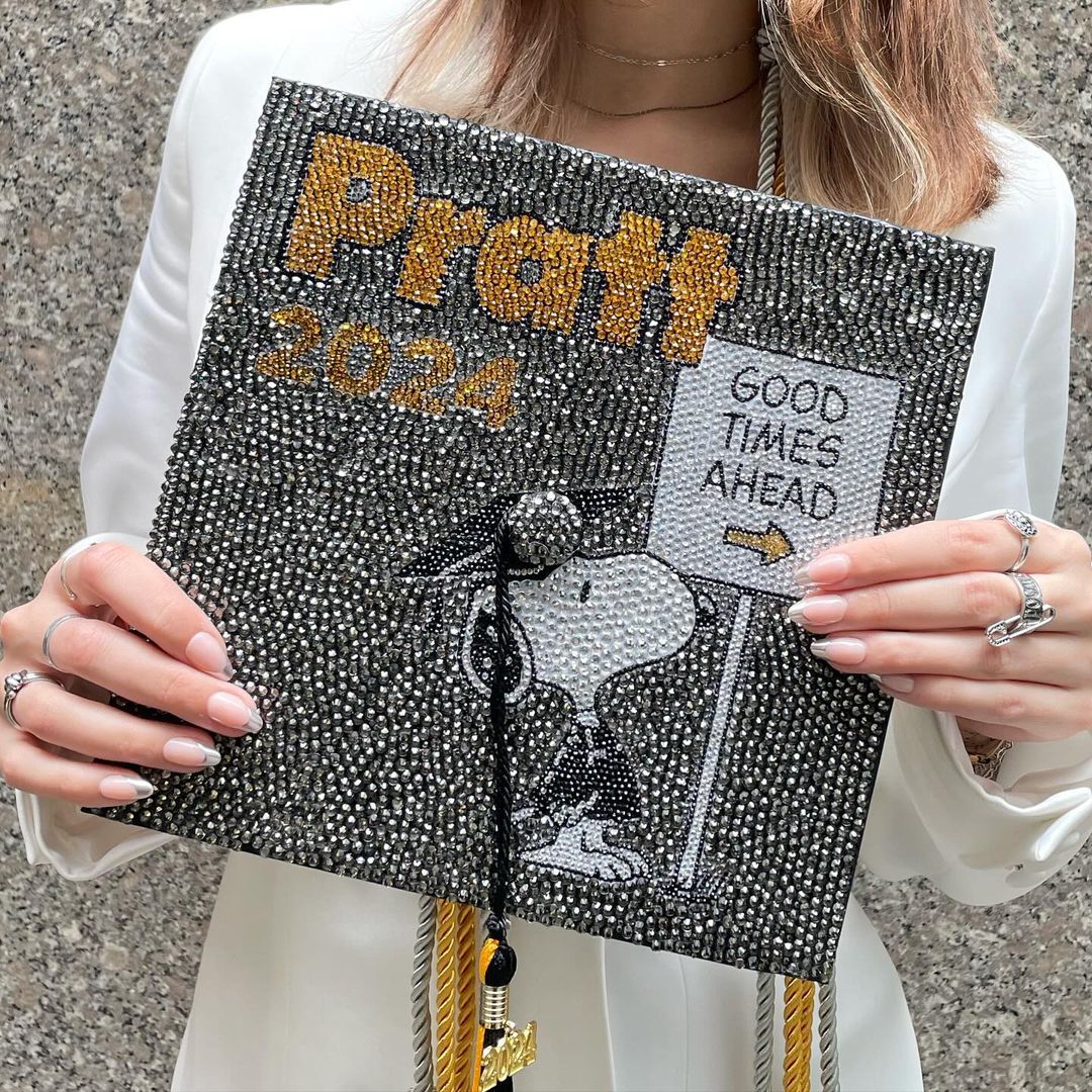 Zoomed-in photo of someone holding a mortarboard bedazzled with jewels, featuring Snoopy in front of a sign that reads 