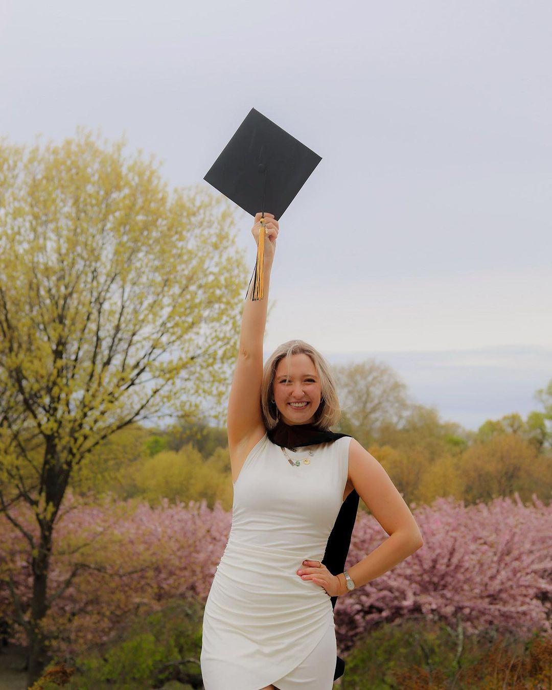 Person in a white dress, wearing a commencement stole, holding up their cap with one hand and smiling in the Brooklyn Botanic Garden.