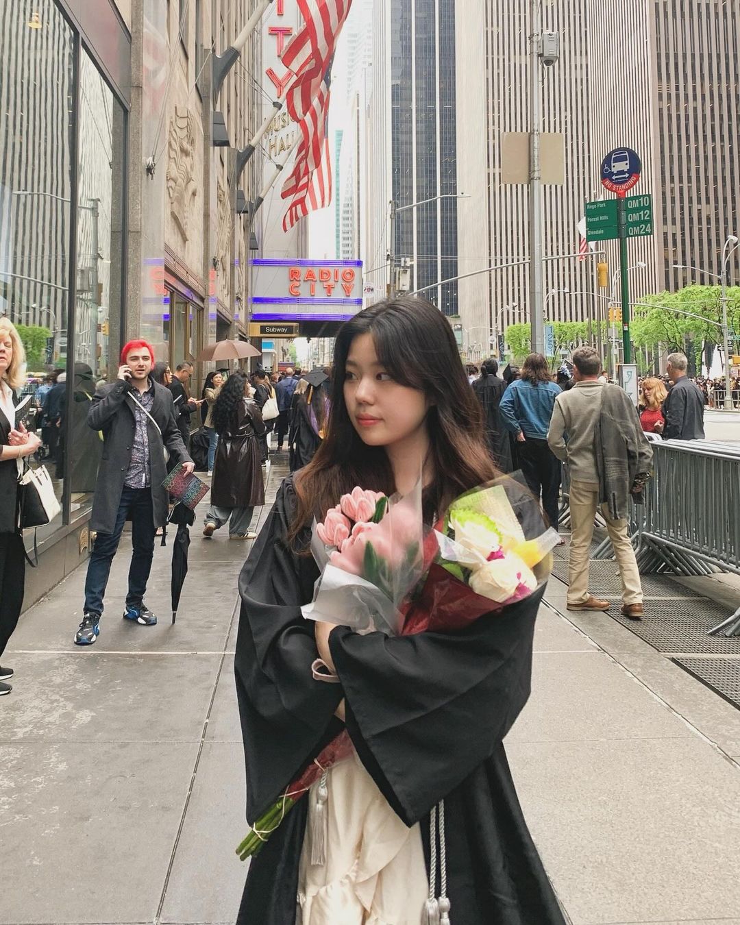 Person standing in a white dress and commencement gown holding flowers, smiling in front of the exterior of Radio City Music Hall.