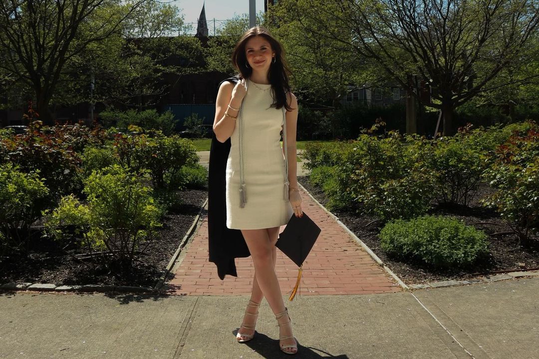 Person in a white dress posing with their commencement gown over their shoulder and their cap in their hand smiling in the Pratt Rose Garden.