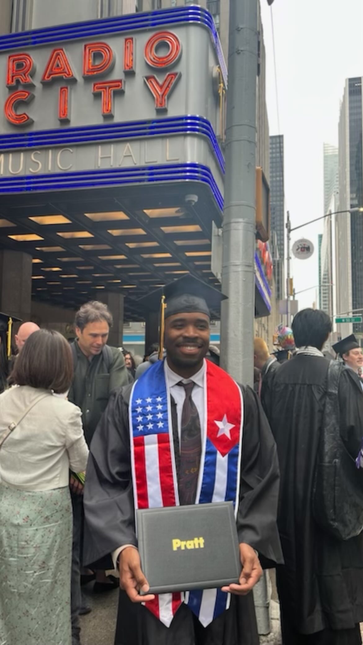 Person standing in their commencement cap and gown smiling and holding up their Pratt diploma in front of the exterior of Radio City Music Hall.