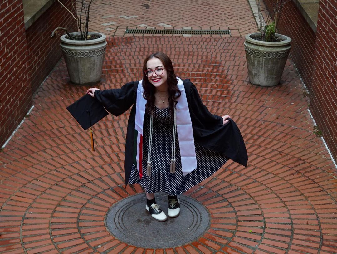 Person in a black and white polka dot dress and their commencement gown curtsying and smiling on Pratt's Brooklyn Campus while holding their commencement cap.