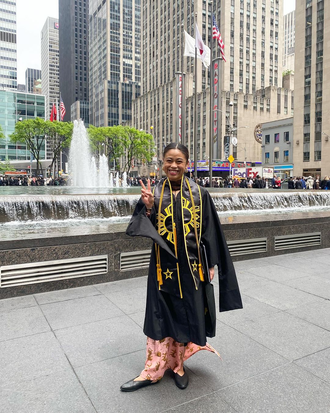 Person in a commencement gown standing and holding up a peace sign in front of the exterior of Radio City Music Hall.