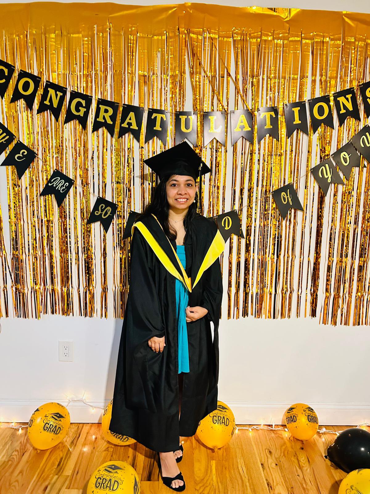 Person in a blue dress and their commencement cap and gown posing and smiling in front of streamers and a banner that says 