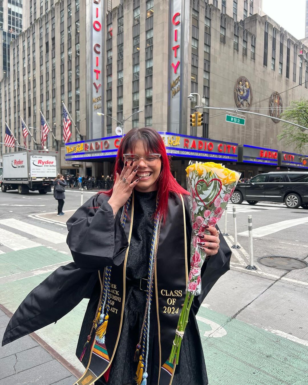 Person in their commencement gown standing in front of the exterior of Radio City Music Hall laughing.