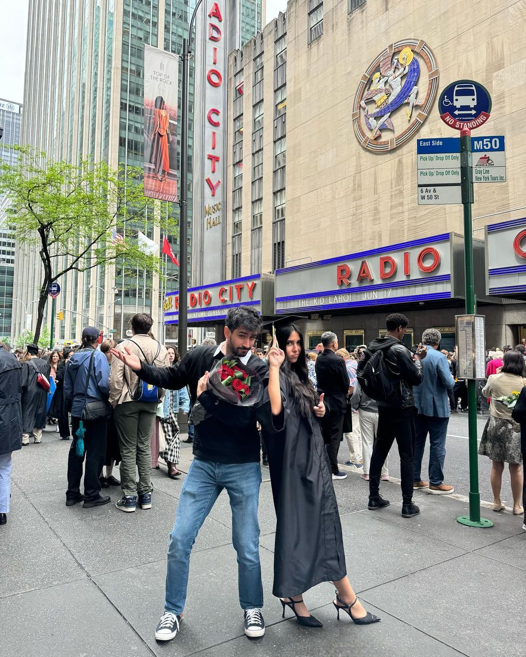 Person in their commencement cap and gown back to back with another person not in commencement attire both posing in front of the exterior of Radio City Music Hall. The person on the left is holding red flowers.