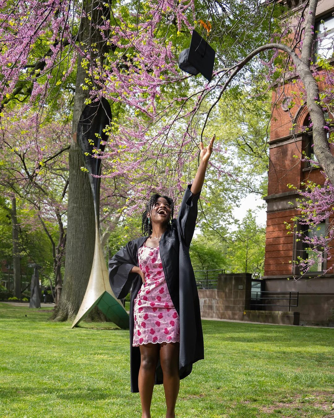Person in their commencement cap and gown back to back with another person not in commencement attire both posing in front of the exterior of Radio City Music Hall. The person on the left is holding red flowers.