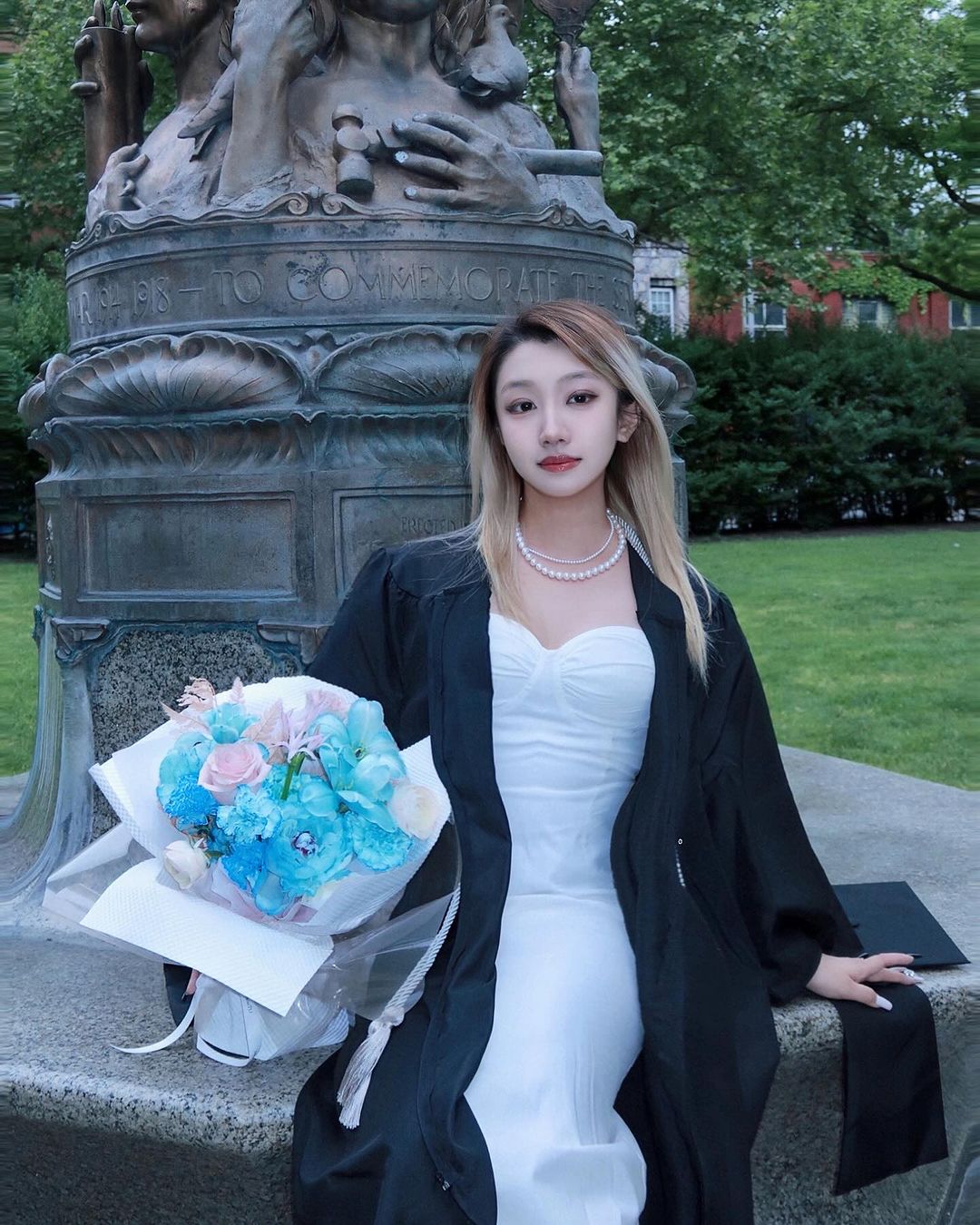 Person in a white dress and their commencement gown sitting in front of a statue smiling and holding pink and blue flowers.