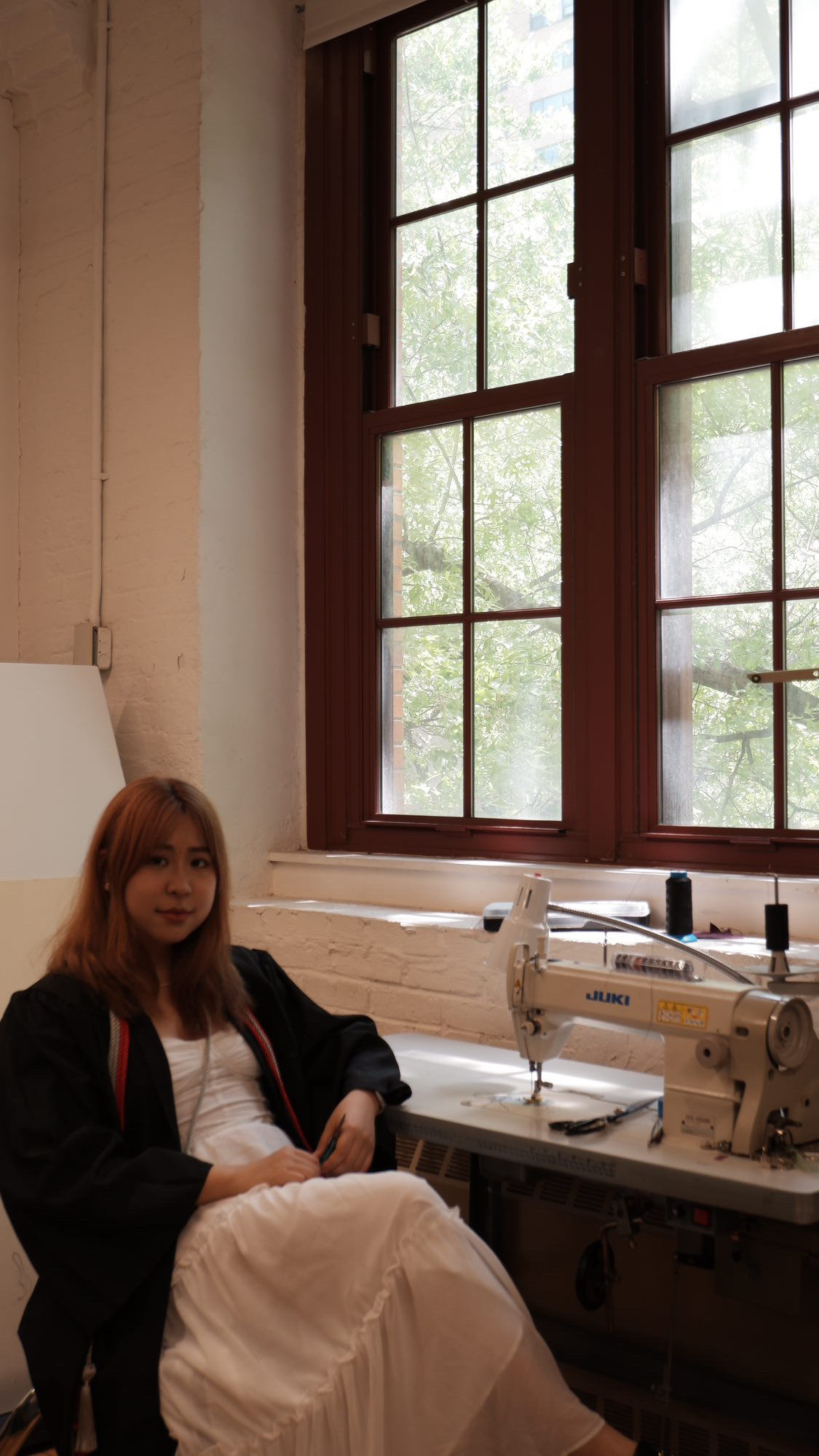 Person in a white dress and commencement cap and gown sitting in front of a sewing machine smiling.