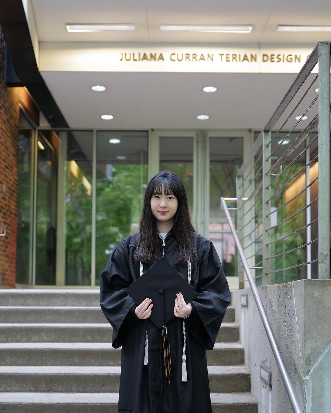 Person standing at the entrance of the Juliana Curran Terian Design Center in their commencement gown holding their cap and smiling.