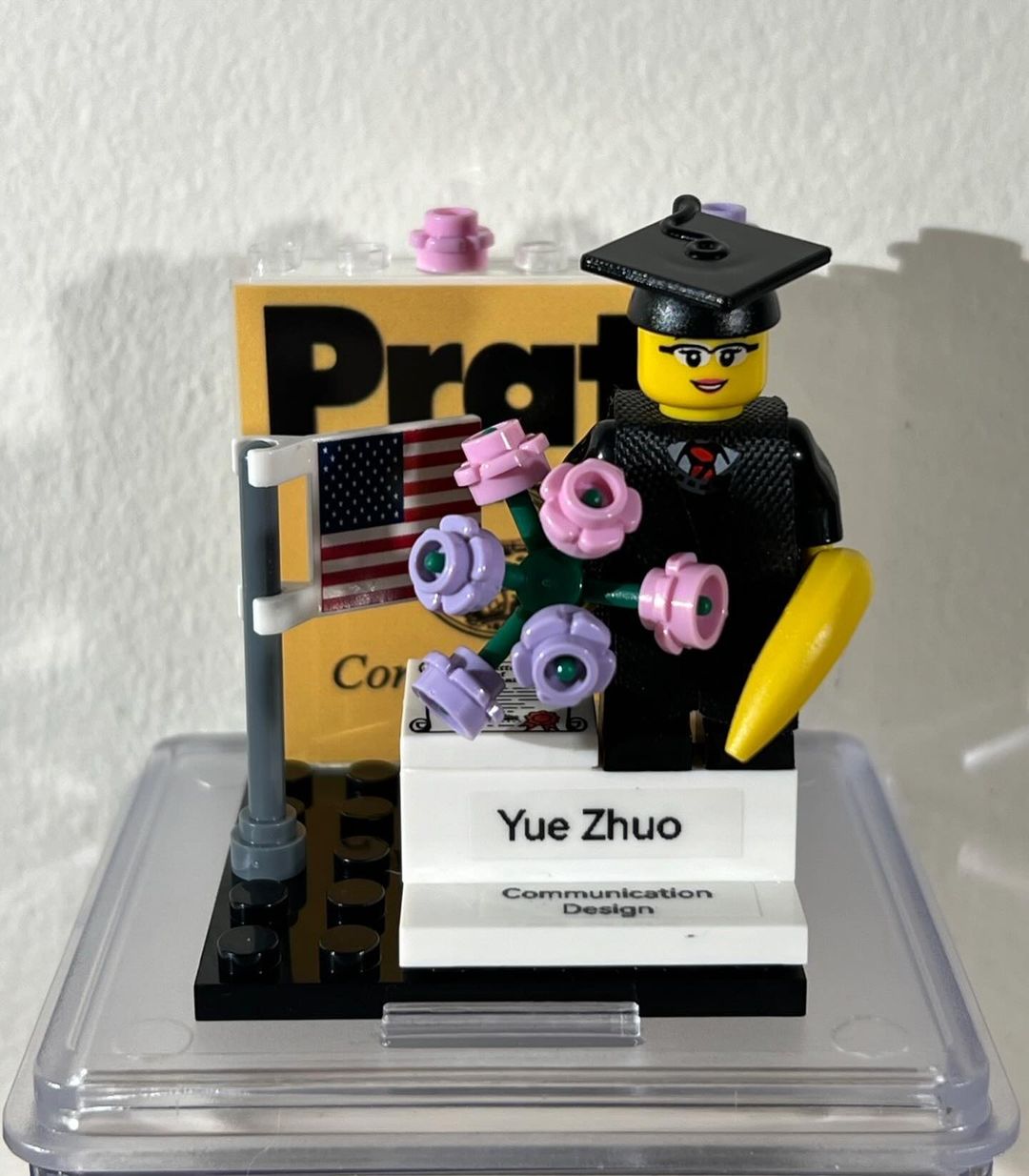 A lego figure smiling in their cap and gown holding flowers on a stand labeled 