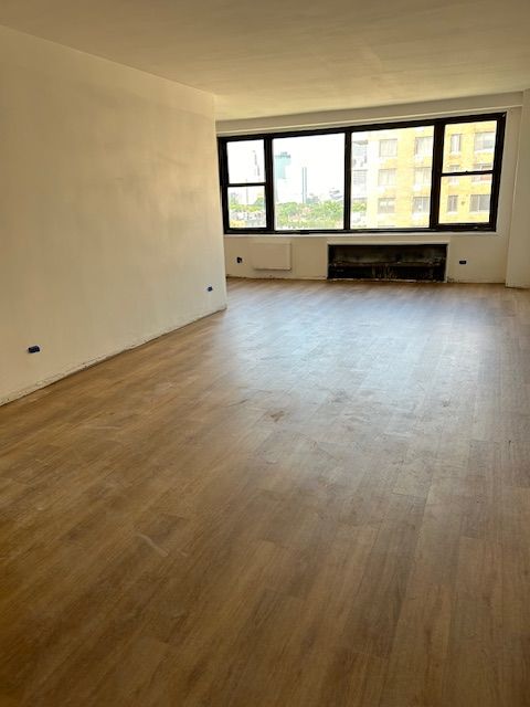 A recently renovated room featuring new wooden flooring, light-colored walls, and a large window at the far end that allows ample natural light to enter. The window offers a view of adjacent buildings, adding an urban element to the space. The room is empty, showcasing the clean and modern look of the renovations, with electrical outlets visible on the walls. The simplicity and openness of the room suggest it's ready for furnishing and personal touches.