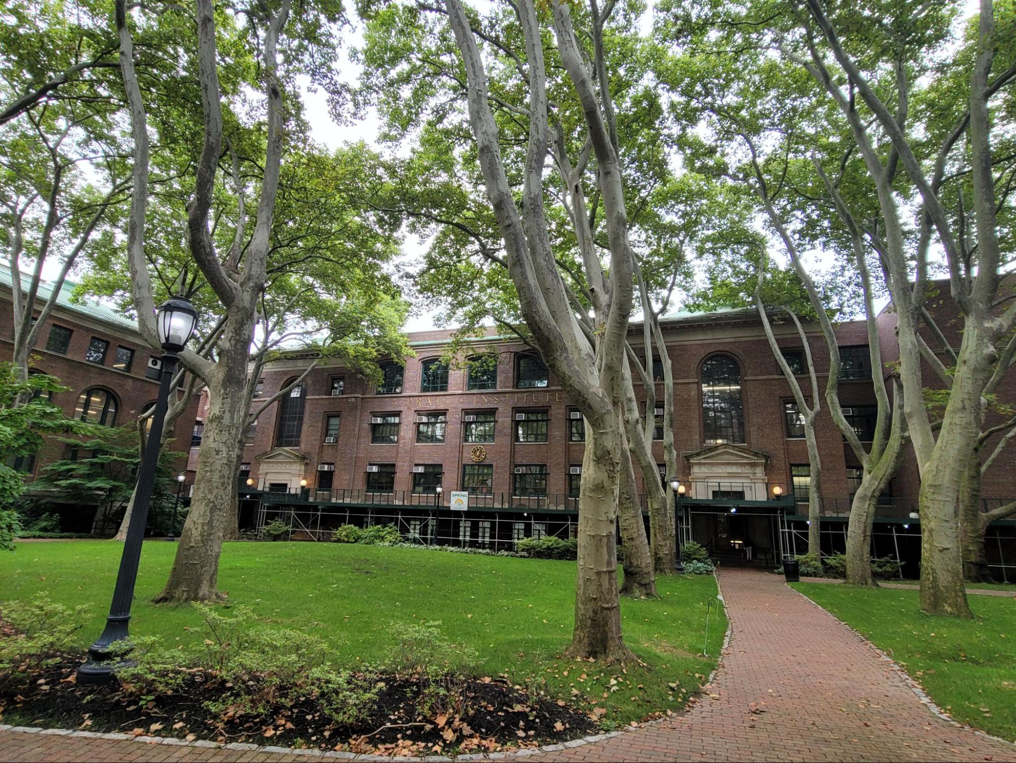 A courtyard in front of a historic, multi-story brick building with large, arched windows and an ornate entrance. The building is framed by mature trees with thick trunks and green foliage, providing ample shade. A well-maintained lawn with a few fallen leaves surrounds a brick pathway that leads to the entrance. A classic black lamppost is visible on the left side, adding a vintage charm to the scene. The overall atmosphere is serene and inviting, typical of a college campus or institutional setting, with an emphasis on blending nature and architecture.