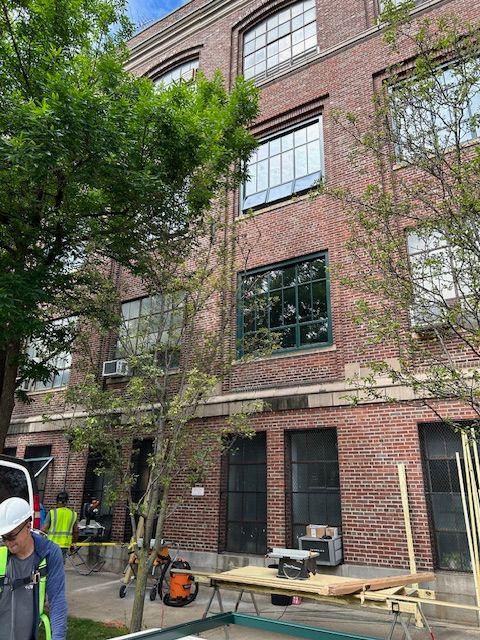 A construction site in front of a multi-story brick building with large windows. The lower part of the building has several tools and materials scattered around, including a workbench with power tools, wooden planks, and construction equipment. A worker wearing a hard hat and safety vest is visible on the left side of the image, focused on his task. Young trees and greenery partially obscure the building, adding a touch of nature to the urban setting. The scene suggests ongoing renovations or maintenance work, with a focus on safety and organization.