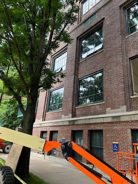 A construction site in front of a multi-story brick building with large windows. The lower part of the building features a concrete base, and there is a piece of heavy equipment with an orange arm visible in the foreground, partially obscuring a young tree. The windows reflect the greenery and the surroundings, creating a blend of urban and natural elements. A blue handicap sign is also visible, indicating accessibility considerations. The overall scene suggests ongoing construction or maintenance work, with safety measures and equipment in place.