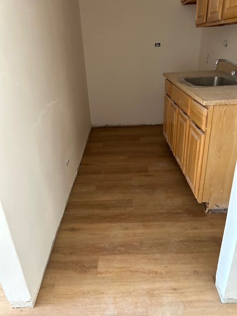 A small kitchenette area with new wooden flooring and plain light-colored walls. On the right side, there is a countertop with a built-in sink and wooden cabinets both above and below the counter. The space appears narrow and minimalistic, suitable for basic kitchen functions. The walls and flooring are recently installed, and the area seems ready for final touches and appliances to be added.