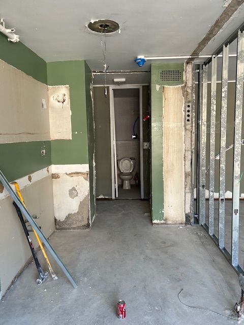 A construction area showing the interior of a partially renovated space. The floor is bare concrete, and the walls have patches of exposed drywall and unfinished sections. There are green and white sections on the walls, indicating an undercoat or preparation for painting. In the center of the image, there is a narrow hallway leading to a small bathroom with a visible toilet. On the right side, metal studs are visible, suggesting ongoing framing work. Various construction materials and tools are scattered around, indicating active renovation work.