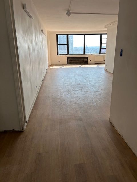An empty room with freshly installed wooden flooring, featuring a large window at the far end that allows natural light to flood in. The walls are freshly painted in a light color, and the space is devoid of furniture or decorations, suggesting recent renovation or preparation for occupancy. The room appears spacious and open, with the window offering a view of the surrounding buildings. The overall appearance is clean and modern, ready for new tenants or further interior design.