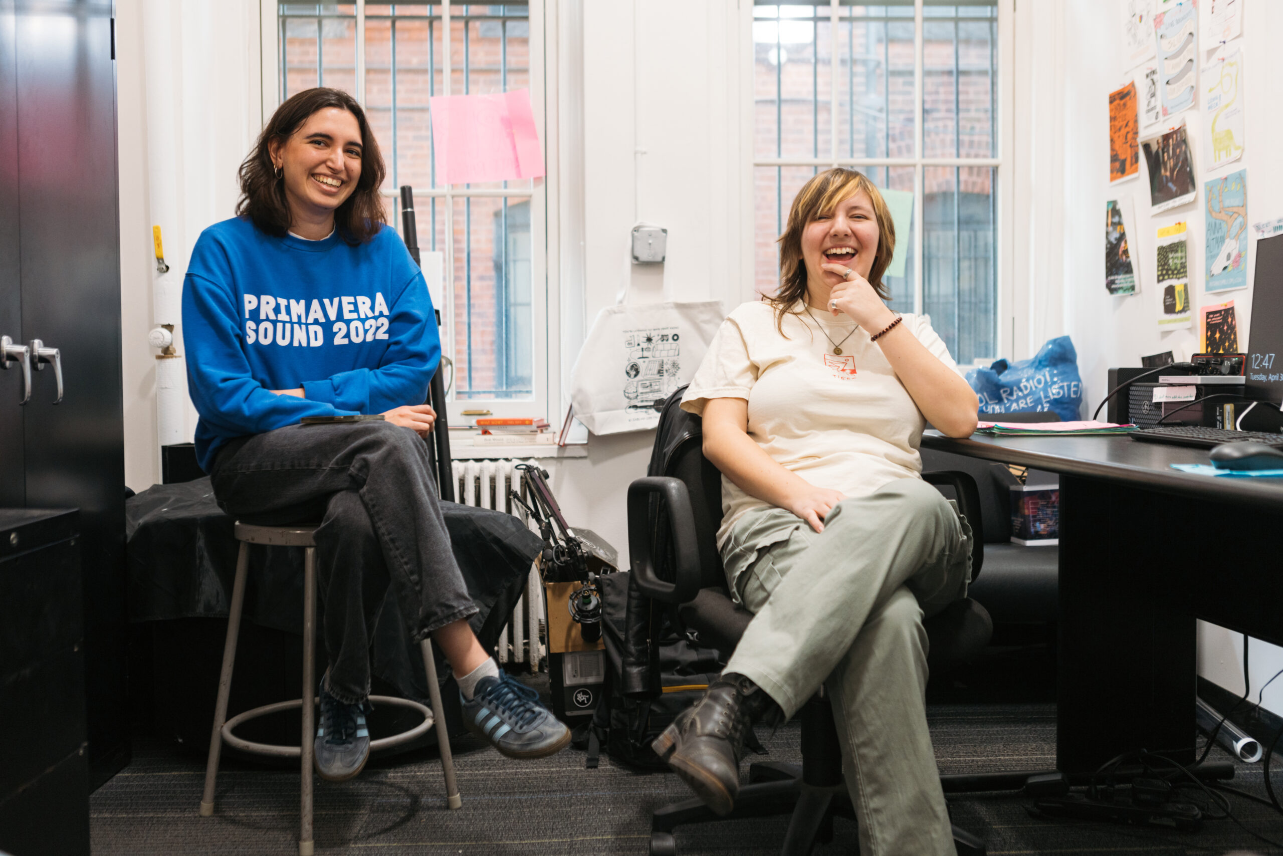 Two people in a radio station laughing. One is sitting in a swivel chair while the other sits on a stool