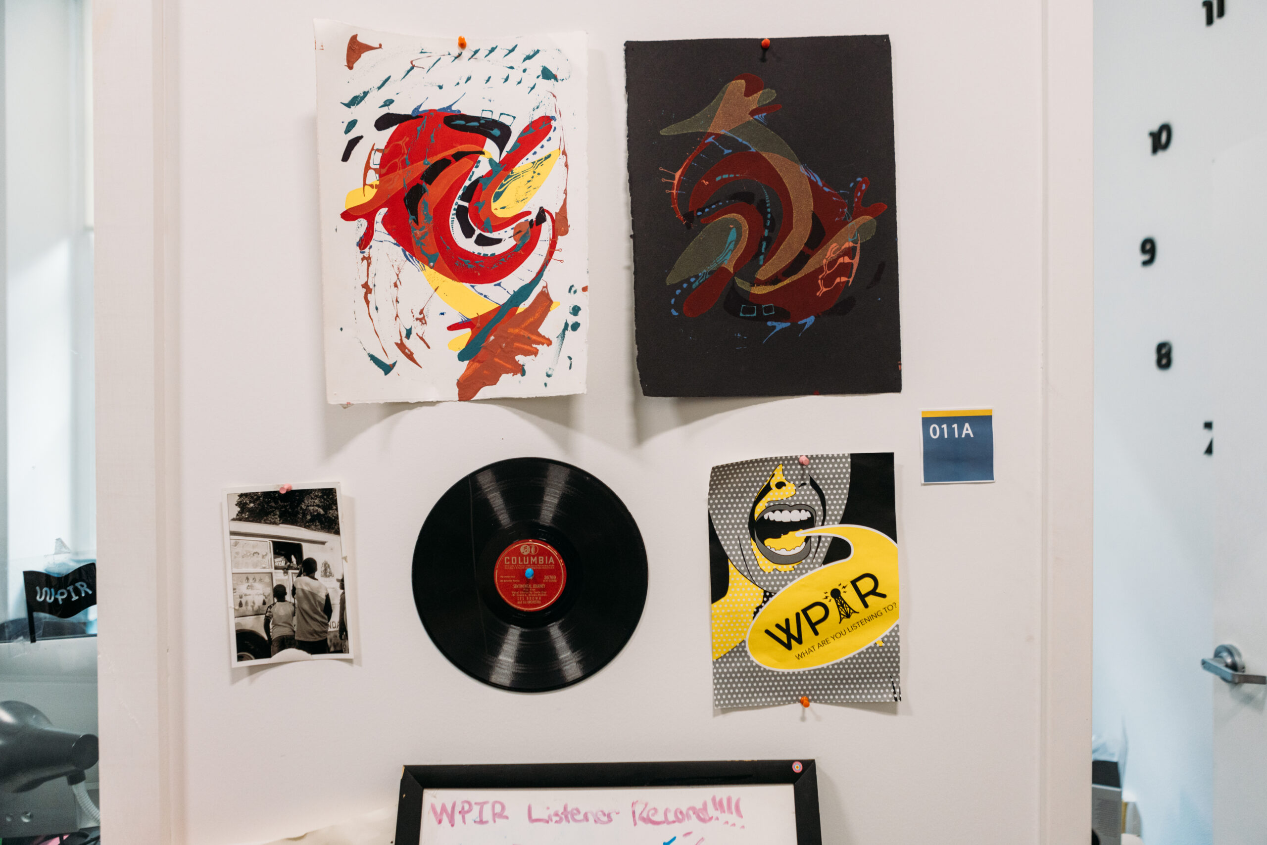 A white wall in a studio with various ephemera on the walls, including a vinyl record, a WPIR poster, and an abstract illustration of a red fish