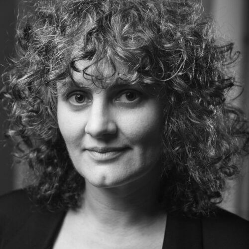 Black and white portrait of a woman with voluminous curly hair, wearing a dark blazer over a light top, looking at the camera with a slight smile and intense eyes.