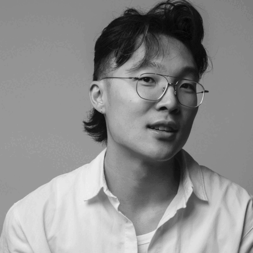 Black-and-white studio portrait of a person with short, layered hair and an undercut, wearing round wire-frame glasses and a light-colored button-up shirt over a T-shirt. The subject is shown from the shoulders up against a plain background, looking slightly off-camera with a calm, attentive expression. Soft, even lighting highlights facial features and creates a neutral, professional tone.