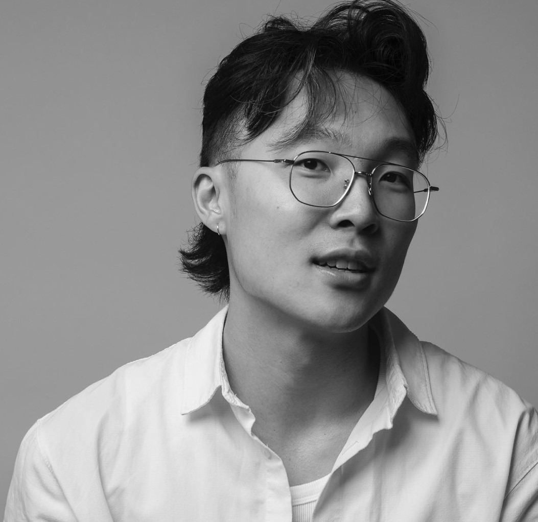 Black-and-white studio portrait of a person with short, layered hair and an undercut, wearing round wire-frame glasses and a light-colored button-up shirt over a T-shirt. The subject is shown from the shoulders up against a plain background, looking slightly off-camera with a calm, attentive expression. Soft, even lighting highlights facial features and creates a neutral, professional tone.