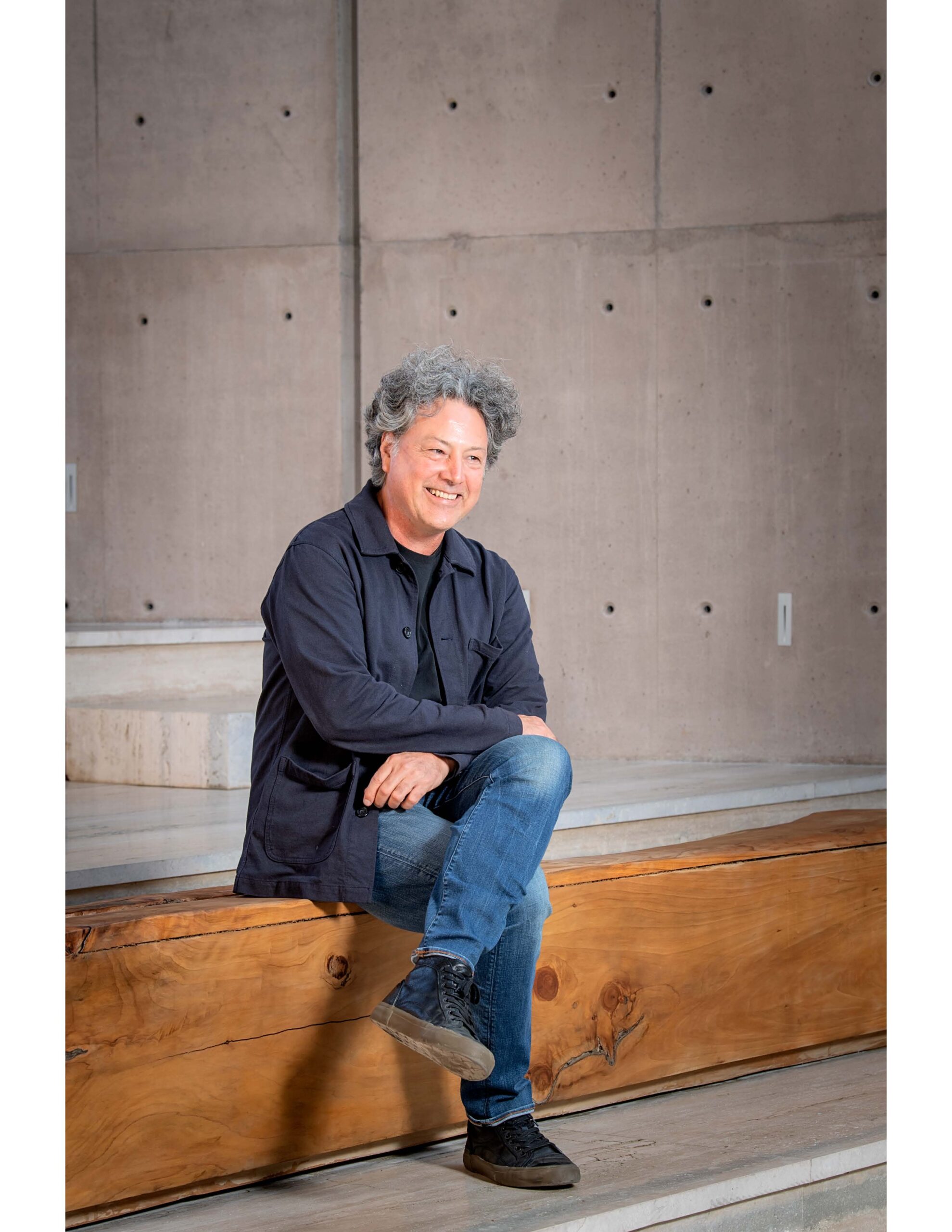 Portrait of a middle-aged man with salt-and-pepper hair, seated on a large wooden beam in front of a concrete wall. He is smiling gently at the camera, dressed casually in a dark button-down shirt and blue jeans, with his hands resting on his knee, exuding a relaxed and friendly demeanor.