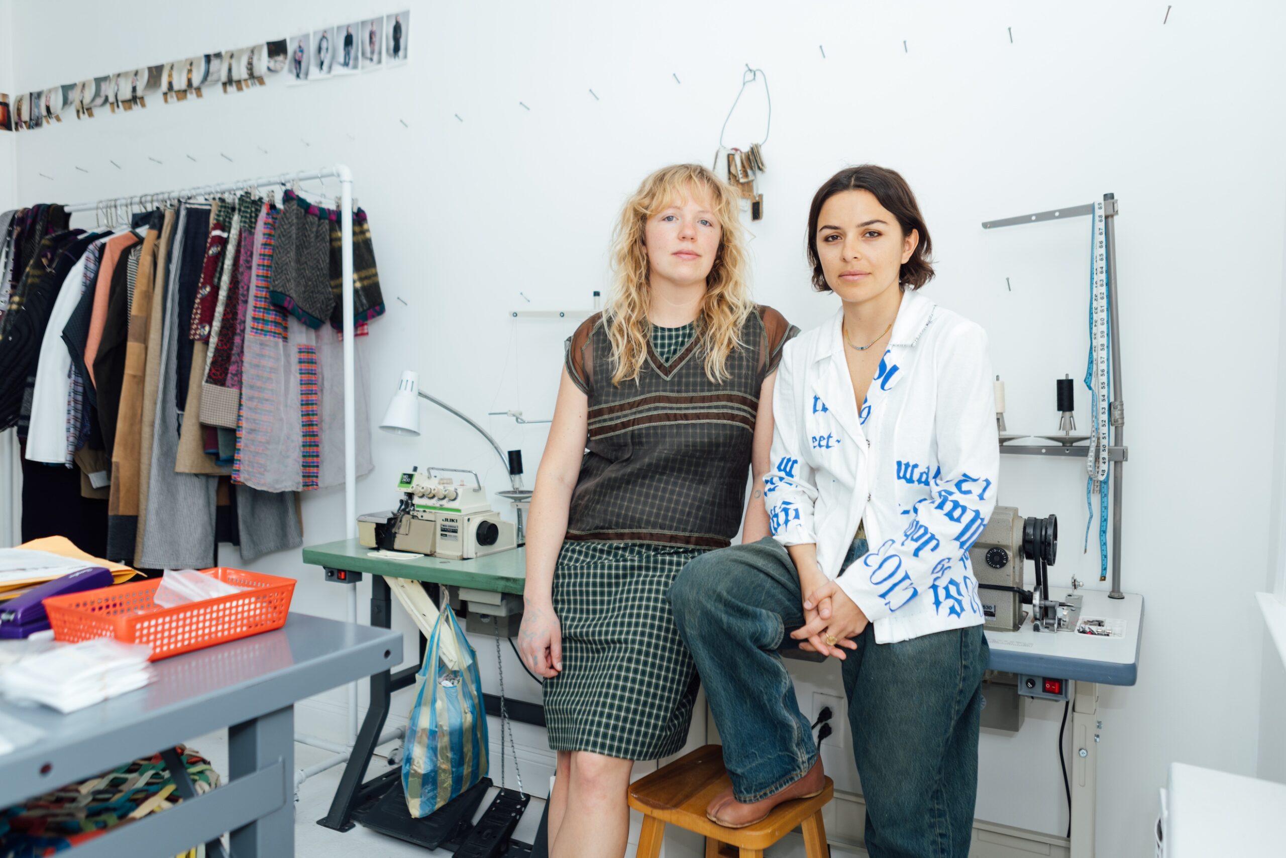 Two people, one in layered sheer and plaid outfit and the other wearing a white suit coat with blue printed lettering on cuffs and sides pose at a studio work table