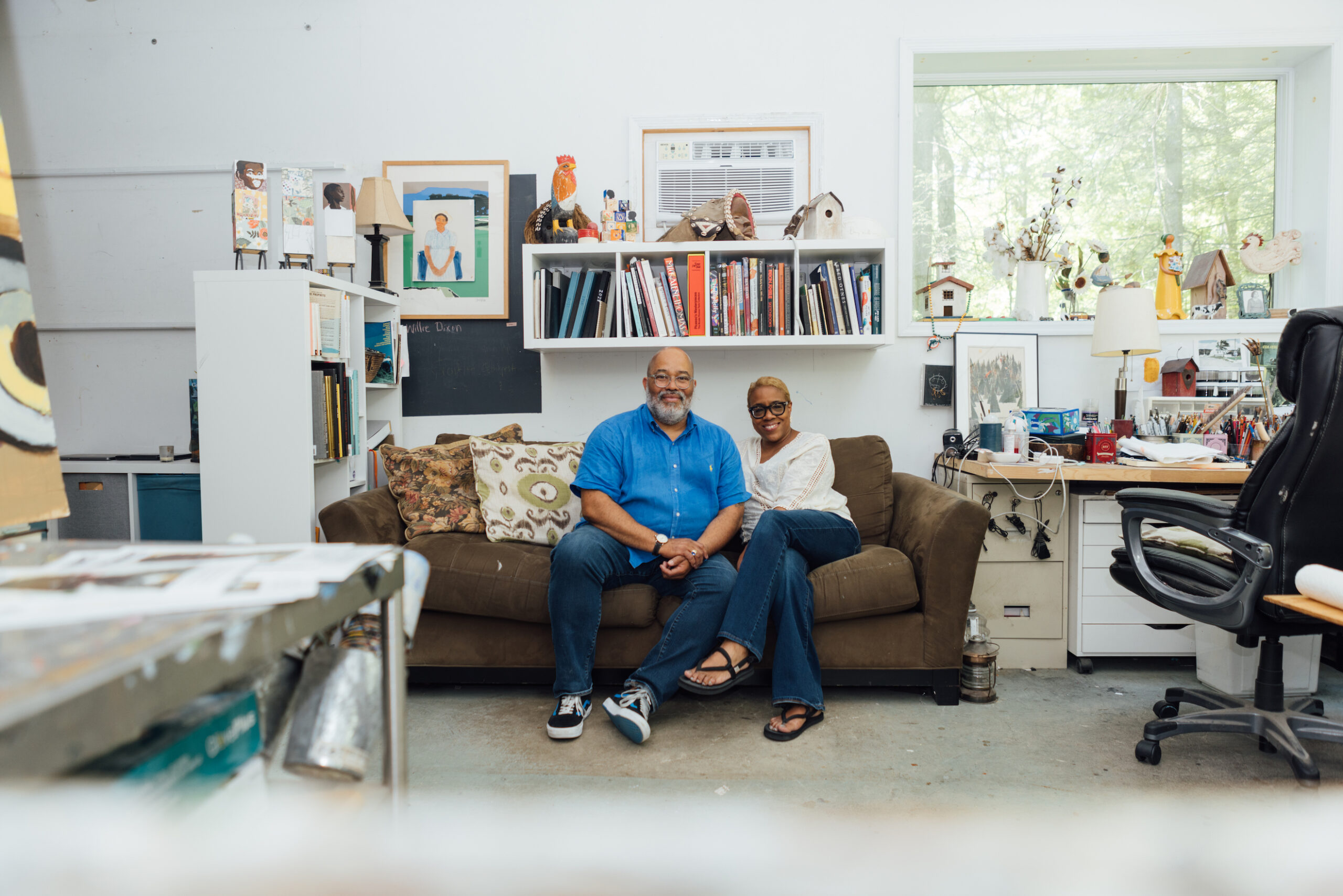 A photograph of two people sitting on a couch in a large, crowded workspace. There is a bookshelf above them and to their right, and a working desk with a window facing the woods to their left.