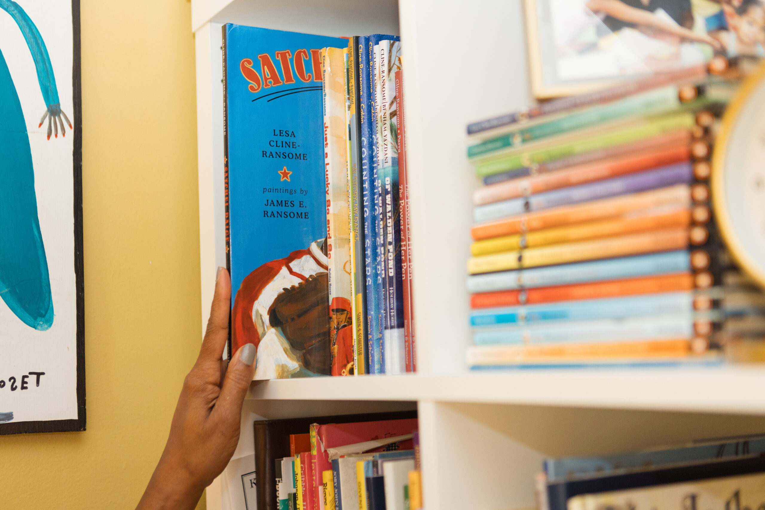 A hand touching a blue picture book peeking out of a white bookshelf with the names “LESA CLINE-RANSOME” and “JAMES E. RANSOME” on display.