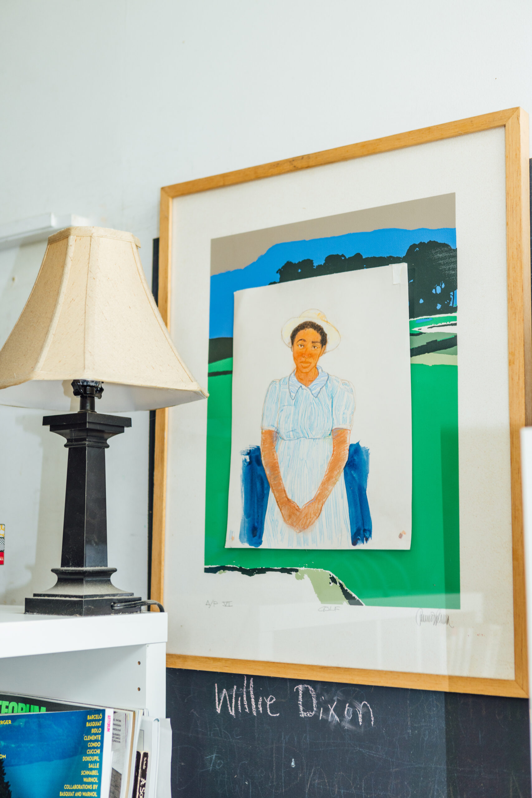 A framed drawing of a person in a light blue dress and a white hat with the name “Willie Dixon” written on a chalkboard below the frame. The image and board are placed behind a bookshelf holding numerous periodicals and a lamp.