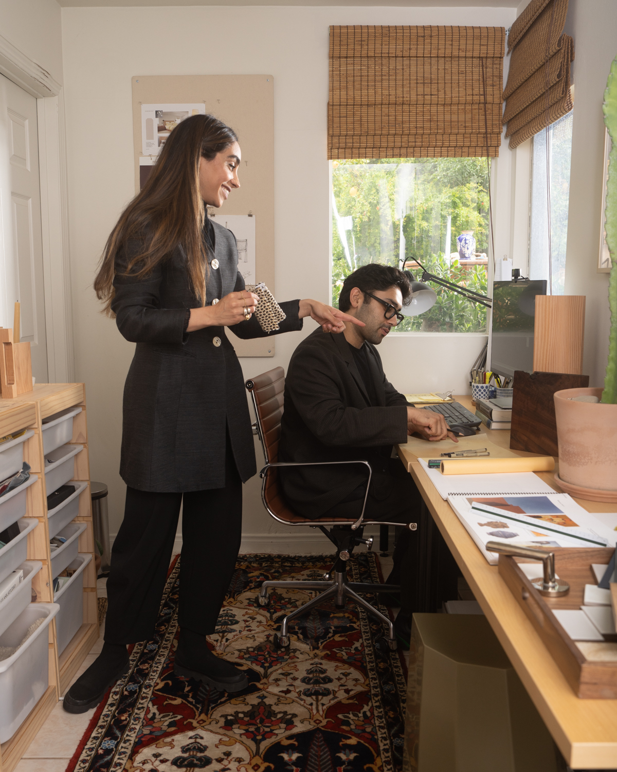 A woman stands smiling and gesturing toward a paper a seated man points at while speaking.