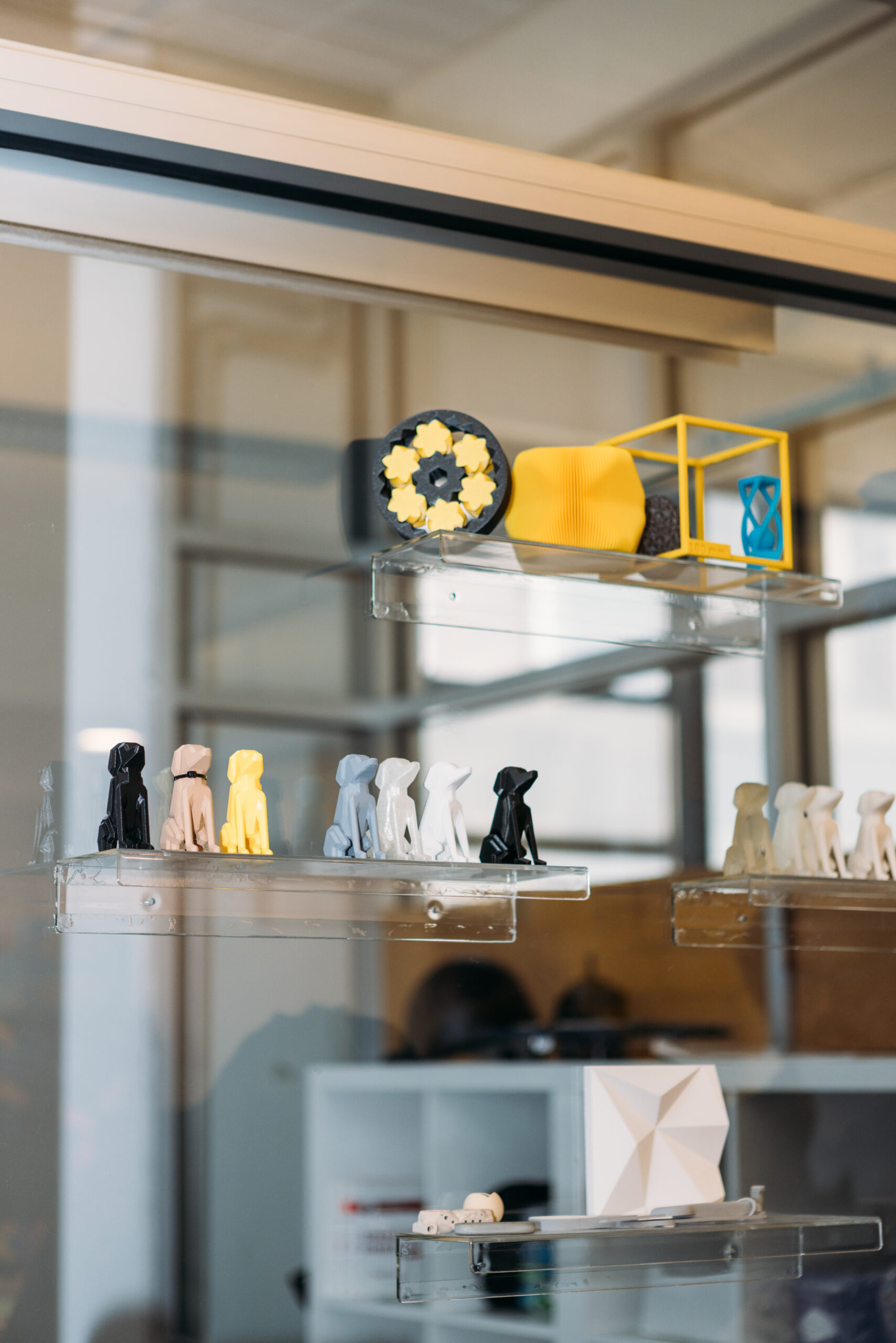 A row of 3D printed figures of dogs and abstract shapes on glass shelves on a glass wall in an office.