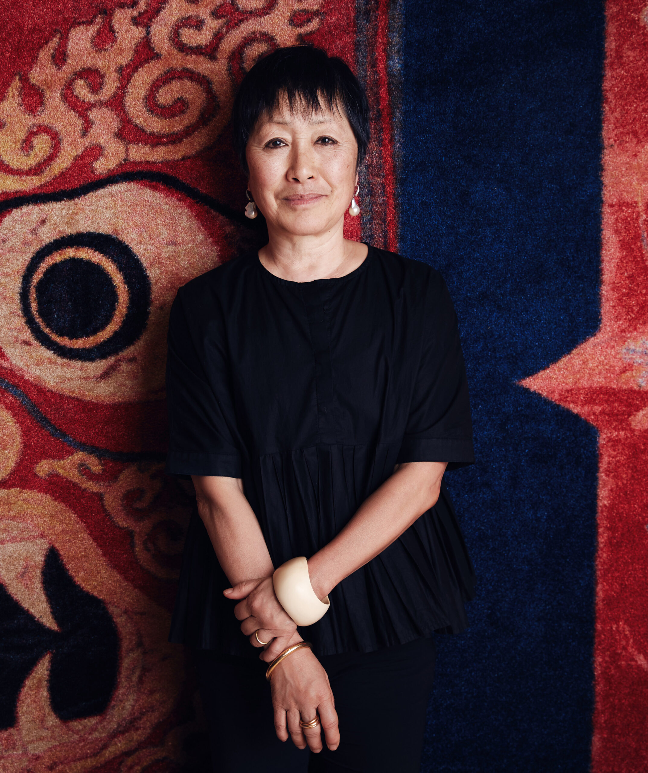 Portrait of a woman with short hair, dressed in a black pleated blouse, standing in front of a vibrant, colorful tapestry with bold designs. She is wearing a white bangle on one wrist and multiple rings, and she gazes calmly at the camera with a gentle expression.