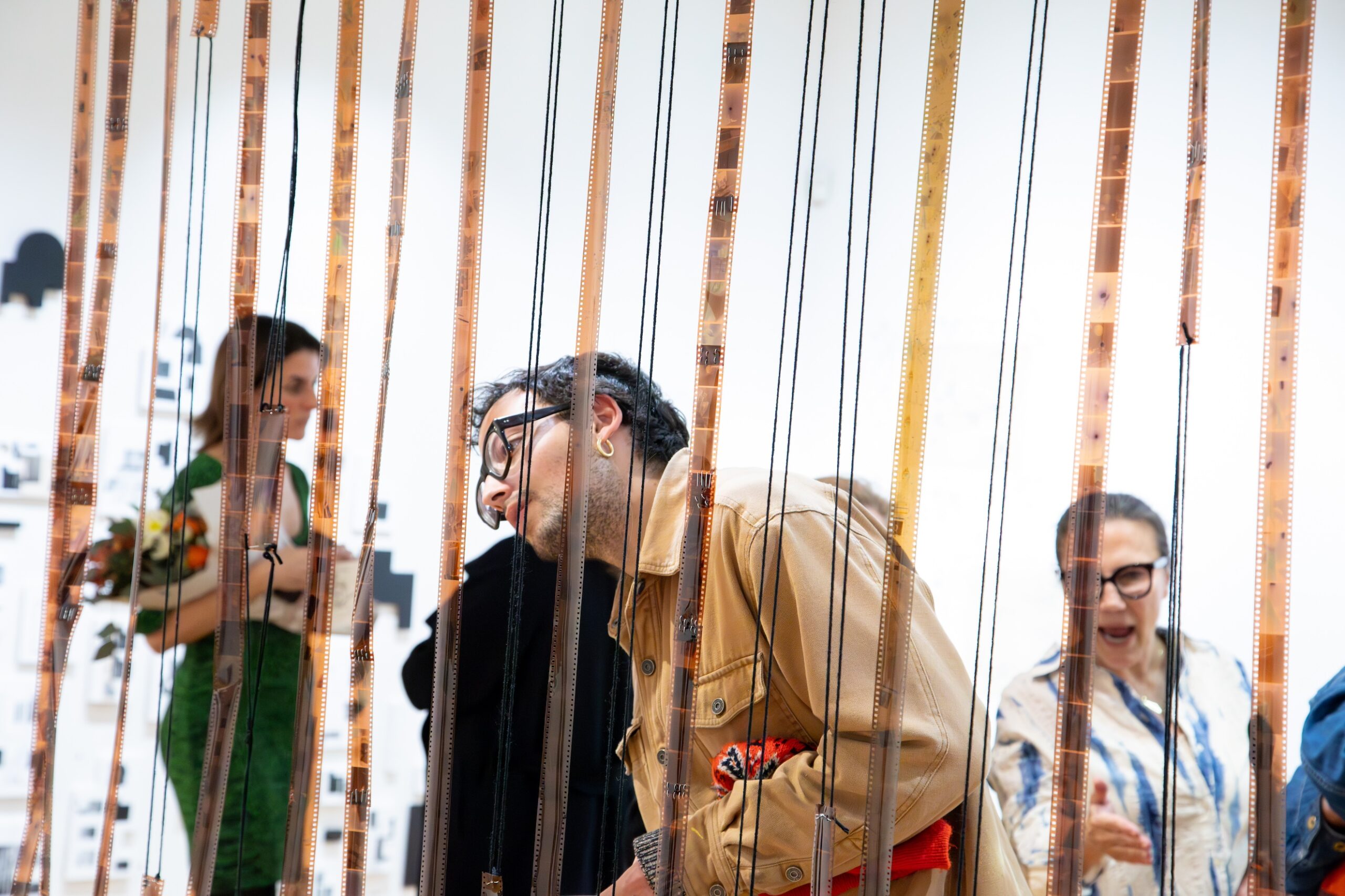 An onlookers gazes at suspended strips of 35mm negatives in a gallery space with white walls; three other visitors are visible in the background