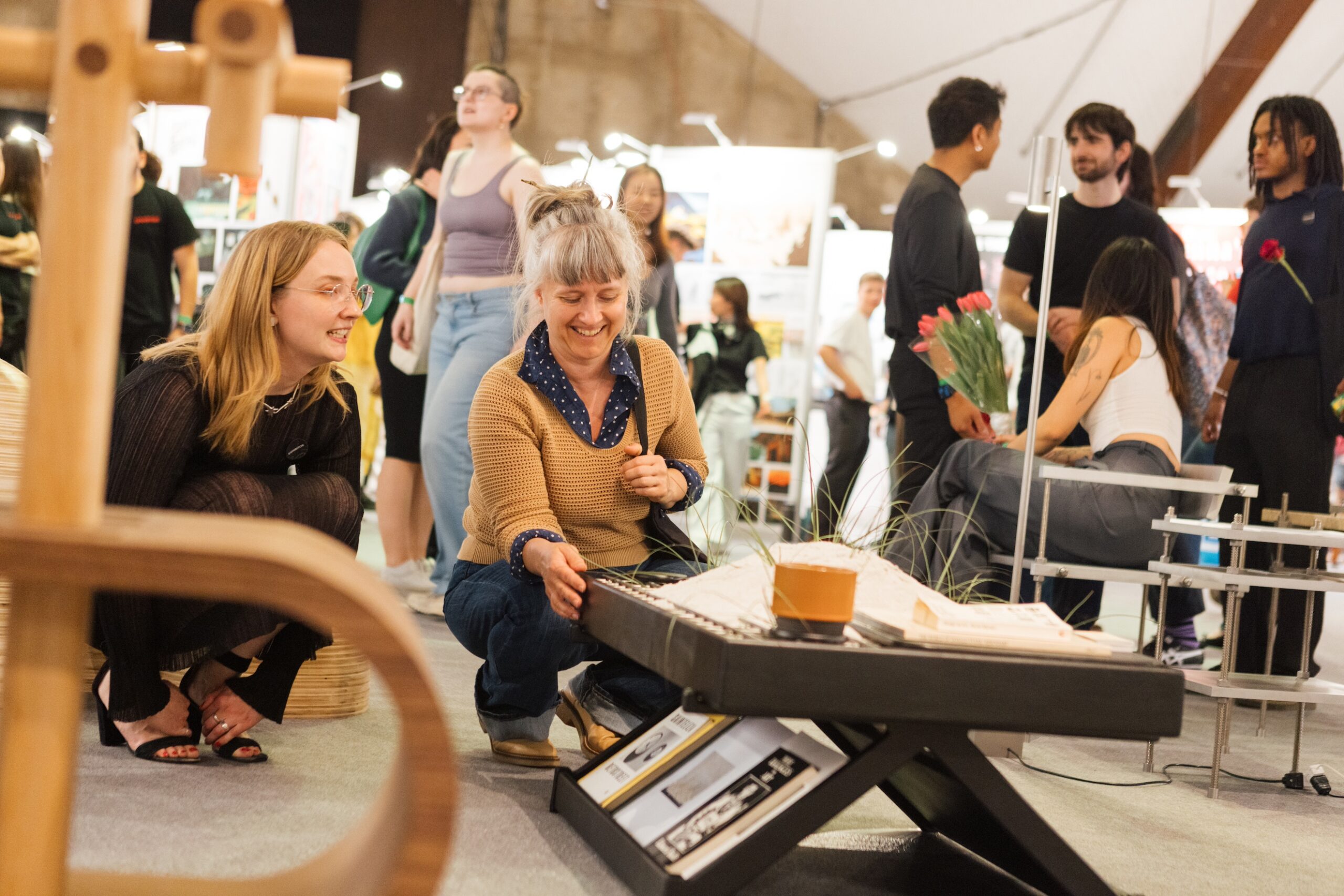 Design exhibition visitors look around at various student furniture designs; in the center of the shot, a person crouches down, smiling, with their hand on a low table