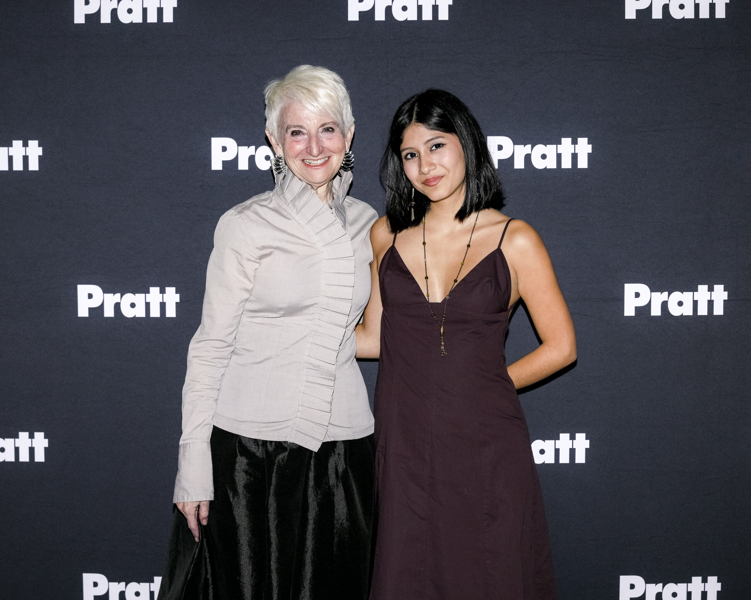 Frances Bronet and Evelyn Meza smile in front of the Pratt photo backdrop.
