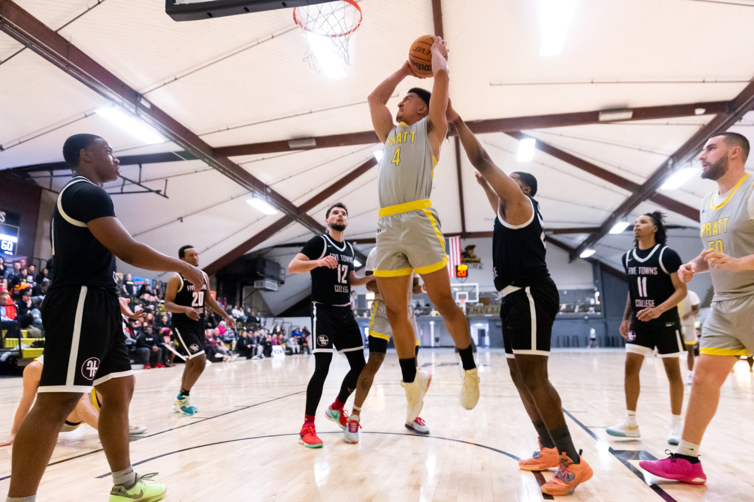 A Pratt basketball player holds the ball and jumps in the air towards the hoop. Around him, the other team is watching or trying to play defense.
