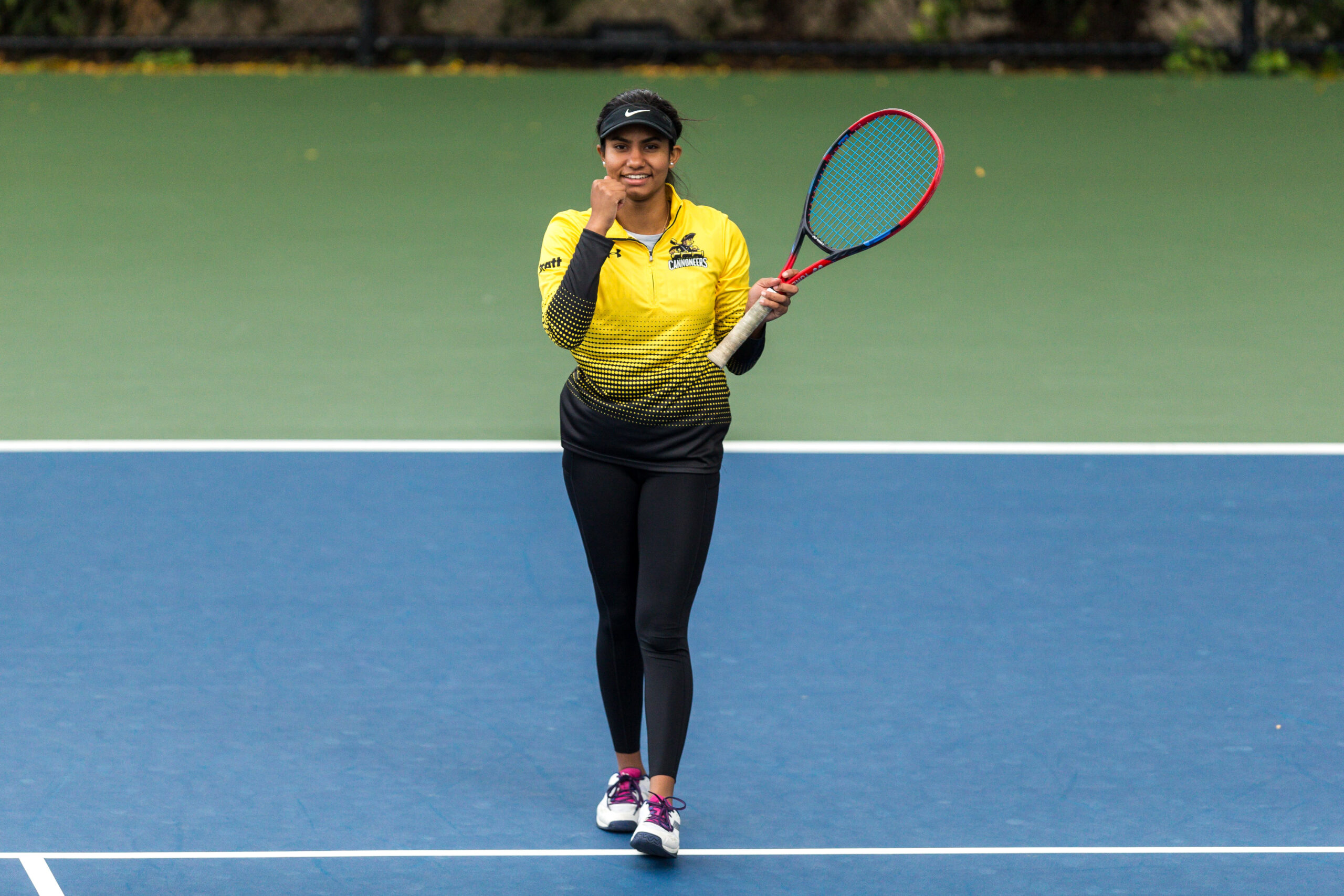 A tennis player smiles on the court. They hold a racquet in one hand and clench the other hand into a fist in front of their face as a sign of victory.