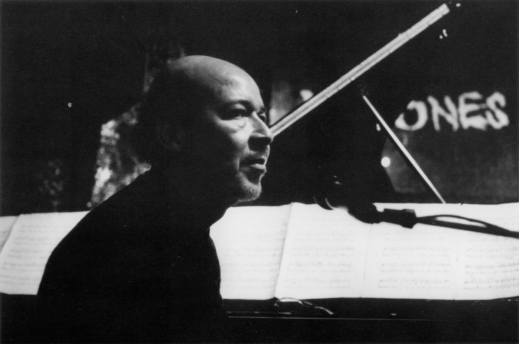 A black and white photograph of a bald man in a black turtleneck sitting behind a microphone. To his left is a piano holding splayed out sheet music.