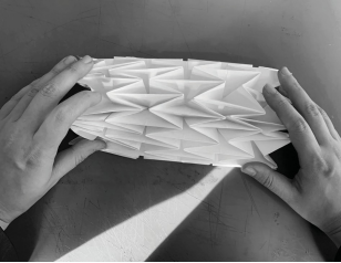 Two hands holding a folded paper structure with geometric, triangular shapes, photographed in black and white.