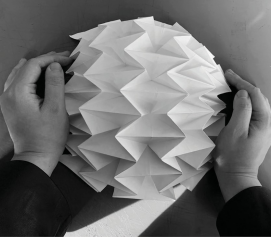 Two hands carefully holding a white, folded paper structure with a textured, star-like surface, showcasing the intricate folds and depth of the design.