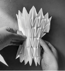 Two hands holding an extended paper structure with sharp, pointed folds, creating a dynamic, layered appearance, photographed in black and white.
