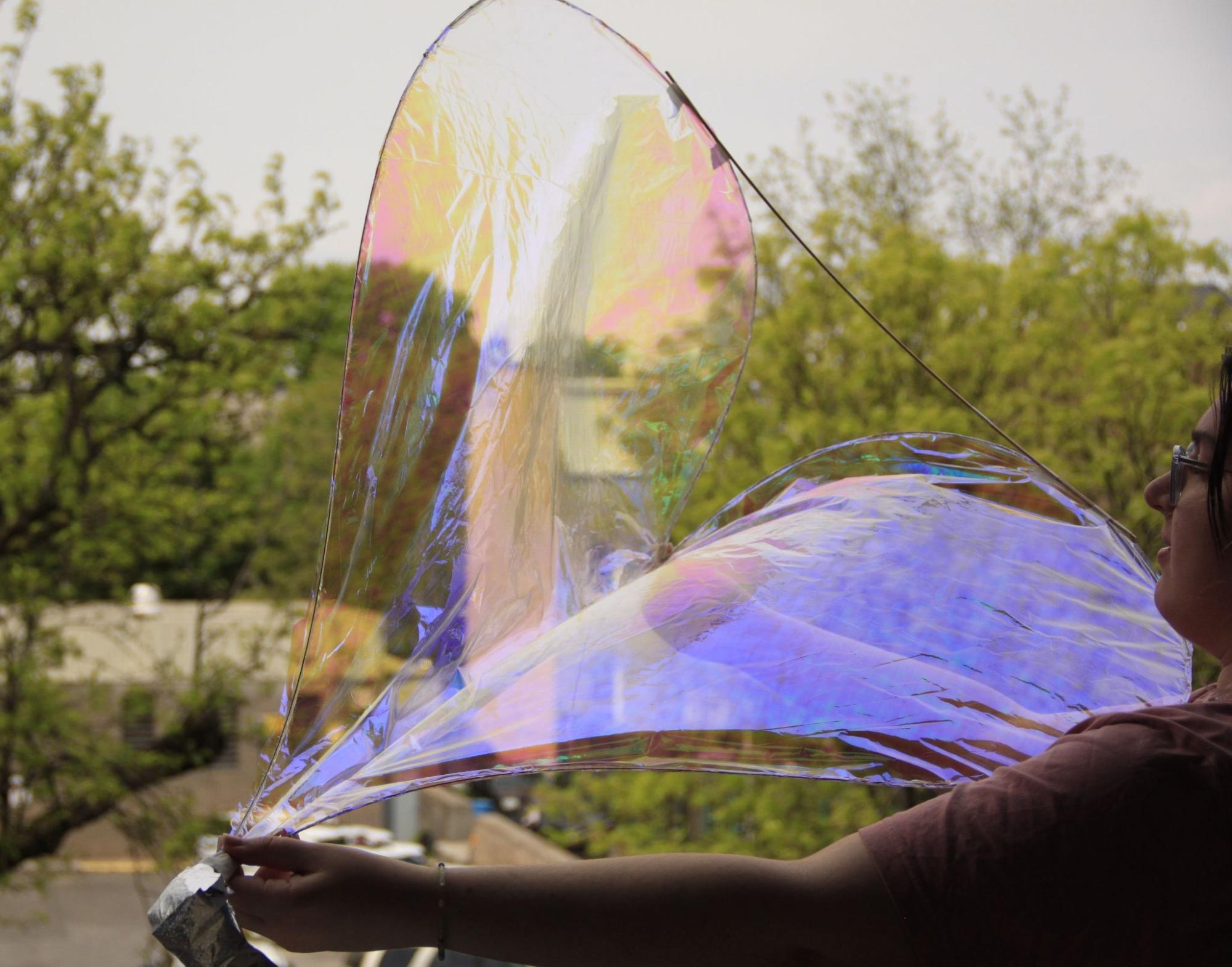Person holding a large, iridescent film that reflects various colors, resembling a bubble, with trees in the background.