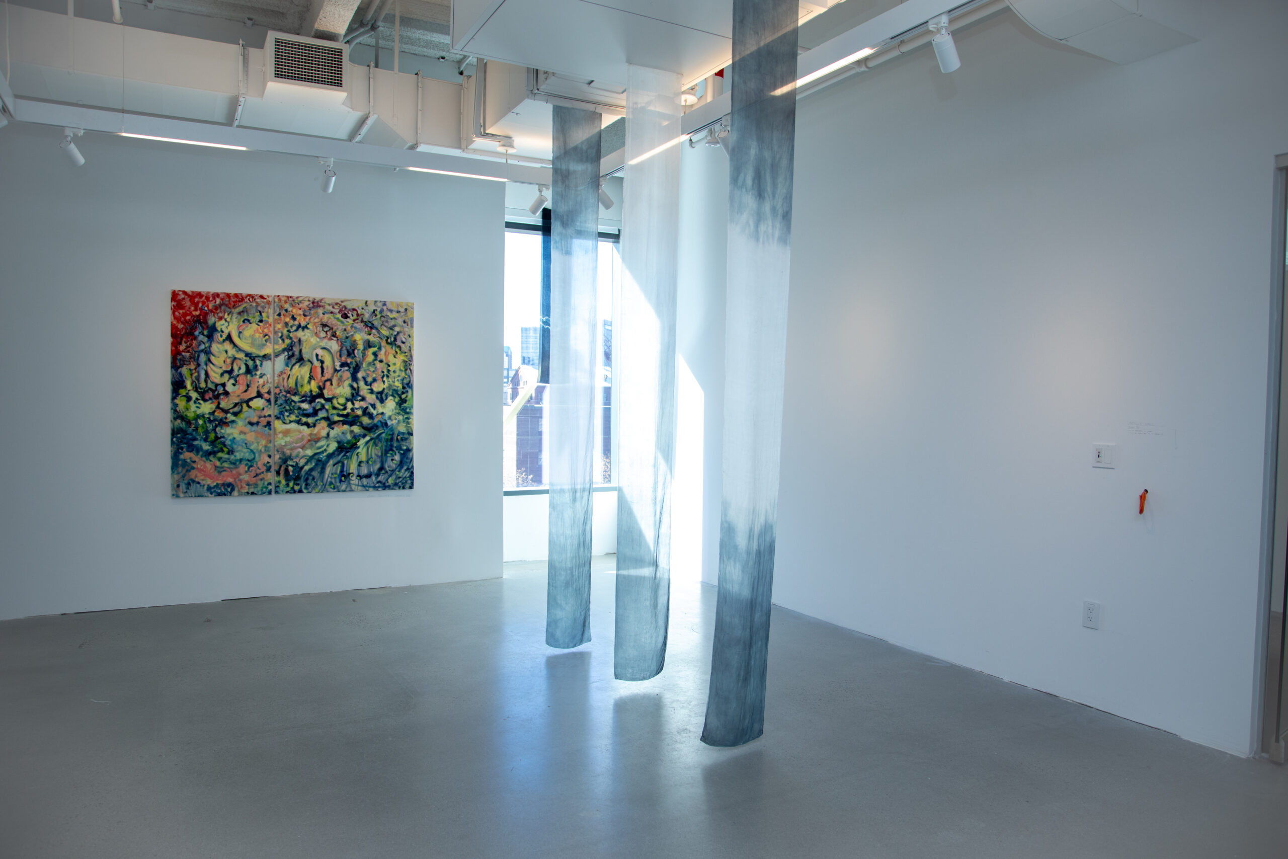 A minimalistic exhibition space with a vibrant, abstract painting on the left wall. In the center of the room, three sheer fabric panels hang from the ceiling. Sunlight streams through the windows.